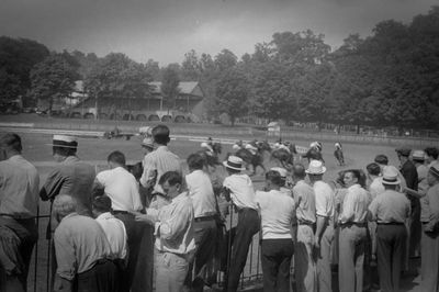 Foto aus den USA, aus dem Jahr 1938, schwarz-weiß: Eine größere Menge Zuschauer betrachtet ein Pferderennen in Lancaster, Ohio. Wir sehen ausschließlich Männer, etwas schräg von hinten fotografiert, aufgereit an einer Balustrade, gekleidert in hellen Sommerhosen und meist weißen Hemden, deren Kragen aufgeknöpft und die Ärmel hochgekrempelt sind, einige tragen Sommerhüte, sogenannte Kreissägen. Im Hintergrund sehen wir das Feld des Pferderennens, mit Jockeys besetzte Pferde auf einer Rennbahn, die gerade an den Zuschauern vorbeigerast sind und sich nun in Richtung Kurve entfernen.