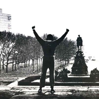 Ein Mann den man von hinten sieht steht in Siegerpose mit ausgebreiteten Armen auf einer Anhöhe. Der Mann schaut auf einen Park mit einem Reiterstandbild, am Rand des Bildes ist ein Hochhaus zu erkennen.