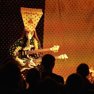 Bild vom Auftritt von Angine de Poitrine. Man sieht eine Person in einer seltsamen gepunkteten Maske mit einem trapezförmigen gepunkteten Hut. Die Person hält eine doppelhalsige Gitarre-E-Bass-Kombination und spielt gerade auf dem unteren Teil des Instruments.