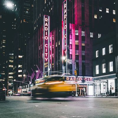 Foto aus dem nächtlichen New York, Straße vor der Radio City Music Hall. Ein Taxi bewegt sich so schnell durchs Bild, dass es wegen der langen Belichtungszeit nur noch schemenhaft erkennbar ist.