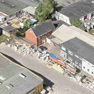 Hinter einer großen Werkshalle steht halb in die Halle hineingebaut ein rot geklinkertes Haus. Rundherum sind andere Industriegeände auf denen alte Stapelkisten, Autos und sogar Boote zu sehen sind, alles aus der Vogelperspektive fotografiert.