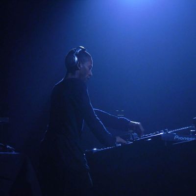 Jeff Mills an seinem Mischpult aus leichter Froschperspektive von hinten rechts fotografiert, in einem in blaues Licht getauchten Raum.