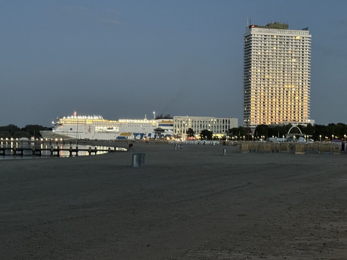 Am Strand von Travemünde, im Vordergrund Strand und Strandkörbe, dahinter in einiger Entfernung die ausfahrende Fähre „Nils Holgersson“, daneben das ungefähr gleich hohe Hotel Arosa und daneben das Maritim-Hochhaus. 