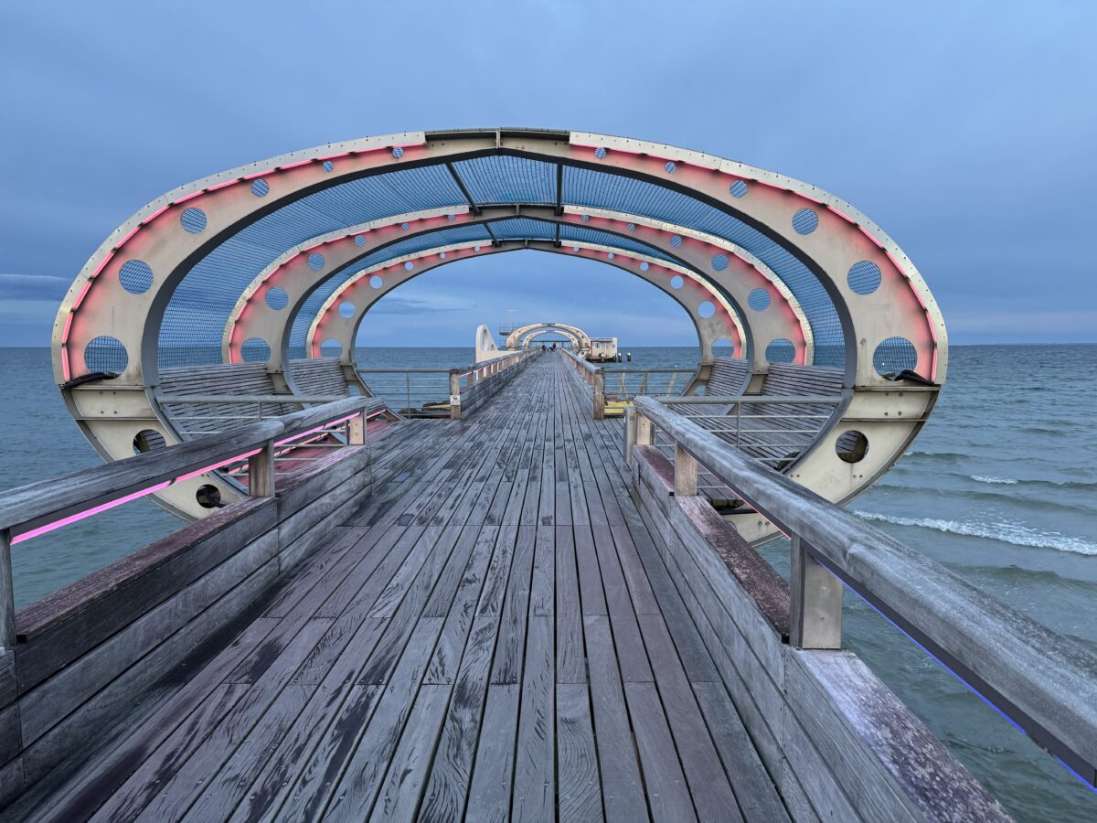 Foto einer Seebrücke in Fluchtpunkt-Perspektive. Der Holzsteg verläuft mittig vom Betrachter weg in den Hintergrund. Links und rechts ist eine Reling aus Holz, darunter das Meer. Im Bildvordergrund ist eine gebogene Überdachung zu sehen, die sich weiter hinten nochmal wiederholt und so einen Tunnelefdwkt erzeugt.
