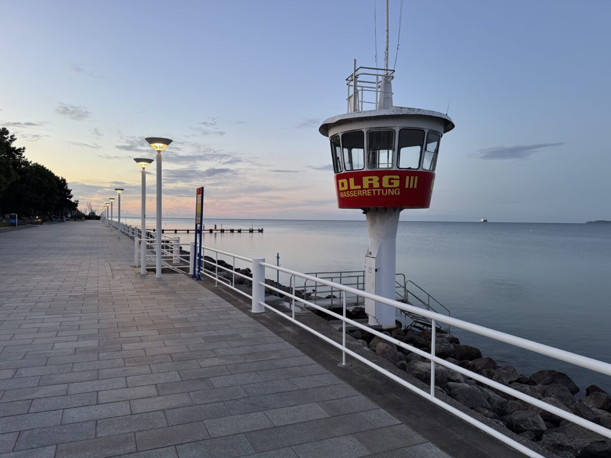 Strandpromenade im Vordergrund, dahinter ein Rettungsturm des DLRG, dahinter die Lübecker Bucht mit rosa Himmel und ein paar Wolken