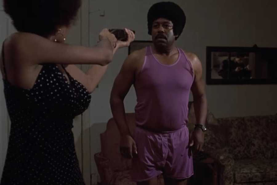 Filmstill: eine schwarze in Kleid bekleidete Frau zielt mit einer abgesägten Schrotflinte auf einen Schwarzen mit Schnauzbart und Afrofrisur, der in rosaner Unterwäsche vor ihr steht.