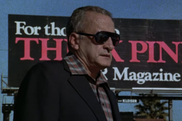 Filmstill aus dem Film Hardcore von 1979. Schauspieler George C. Scott bekleidet mit einem dunklen Sakko, Karohem, einer Pilotensonnenbrille steht vor einem Werbeplakat des Hustler Magazins, dess Aufschrift man aber nicht lesen kann.
