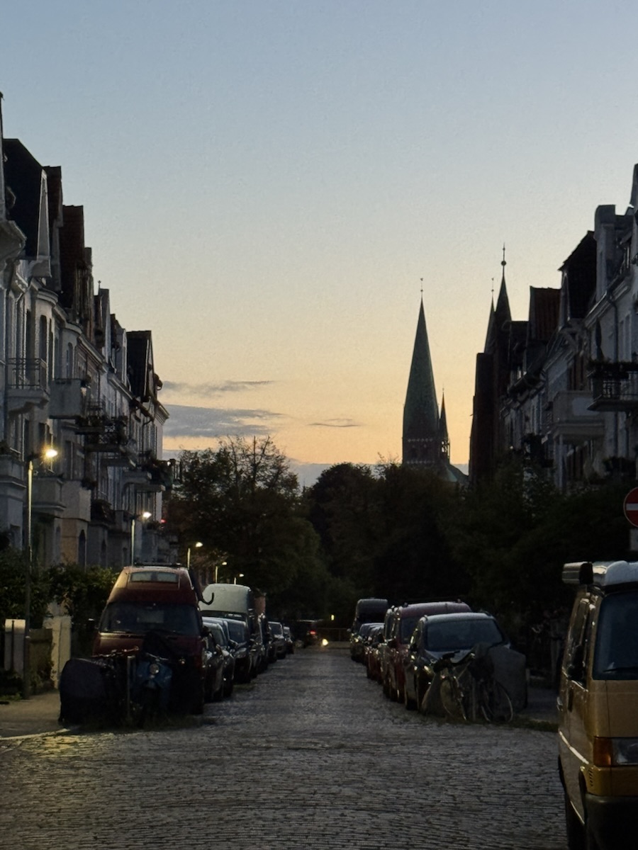 Straßenzug nach Sonnenuntergang, Fluchtpunktperspektive. Mittig Straße mit Altbauten aus den 20er Jahren des letzten Jahrhunderts. Im Hintergrund gegen den Abendhimmel ein Teil der Altstadt-Skyline mit der Marienkirche.