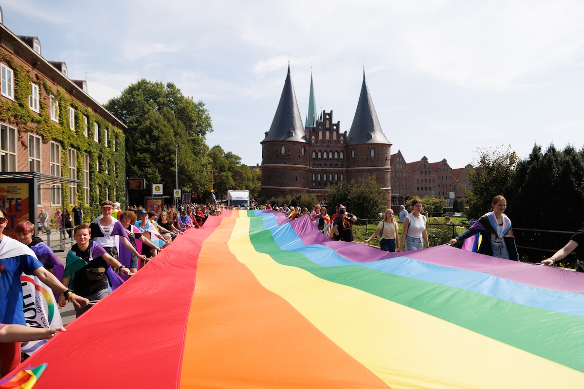 Demonstration CSD vor dem Holstentor, im Bild ist eine riesige Regenbogenfahne, die von vielen Menschen getragen wird und die ganze Straße vor dem Holstentor bedeckt. © Lübecker CSD e.V.