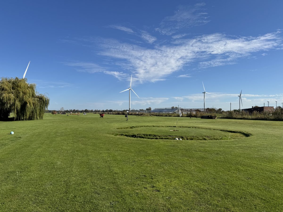 Große Rasenfläche ähnlich eines Golfplatzes, in der Mitte eine kleine Fahne mit einem Graben drumherum, in dem Fußbälle liegen, im Hintergrund Windräder, schönes Wetter, blauer Himmel.